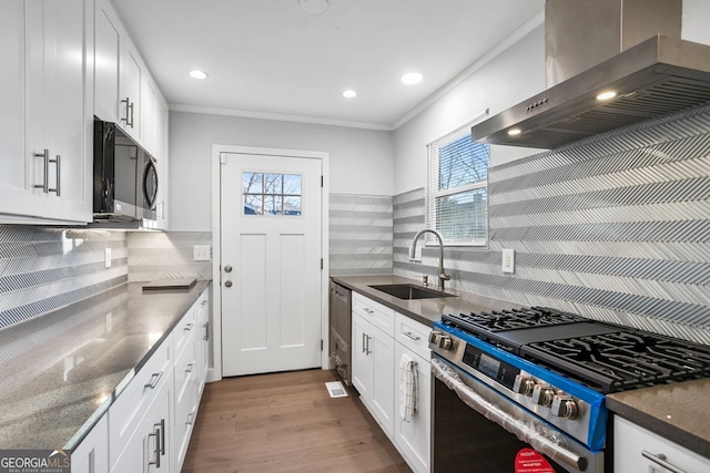 kitchen with appliances with stainless steel finishes, white cabinets, a sink, wood finished floors, and wall chimney exhaust hood