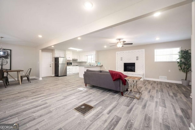 living room featuring ceiling fan, visible vents, baseboards, a brick fireplace, and light wood-type flooring