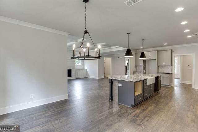 kitchen with light stone counters, pendant lighting, gray cabinetry, ornamental molding, and a kitchen island with sink