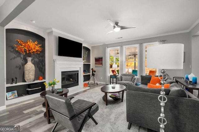 living area featuring wood finished floors, a ceiling fan, baseboards, a glass covered fireplace, and crown molding