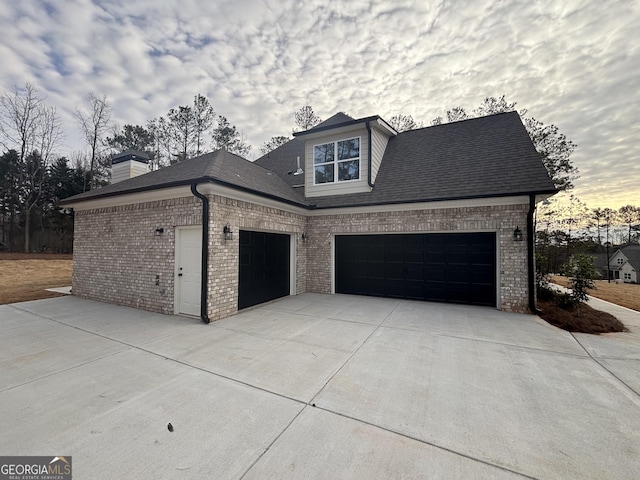 property exterior at dusk featuring a garage, concrete driveway, brick siding, and a shingled roof