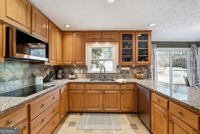 kitchen featuring brown cabinetry, light stone counters, glass insert cabinets, stainless steel appliances, and a sink