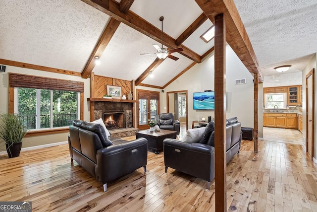 living room featuring beam ceiling, visible vents, light wood-style floors, a textured ceiling, and a stone fireplace