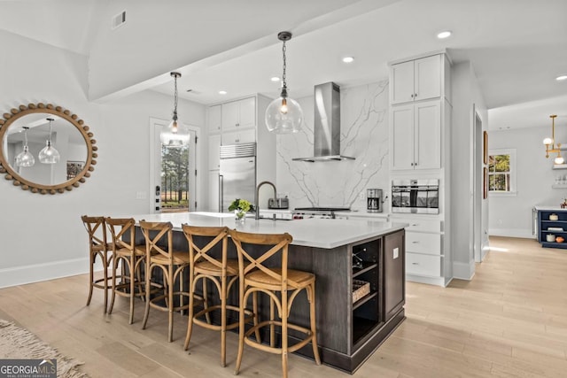 kitchen with light countertops, appliances with stainless steel finishes, white cabinets, wall chimney range hood, and an island with sink