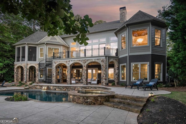 back of property at dusk featuring an outdoor pool, a balcony, stairway, an in ground hot tub, and a patio area