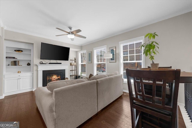 living area with dark wood-style flooring, a glass covered fireplace, a ceiling fan, and crown molding