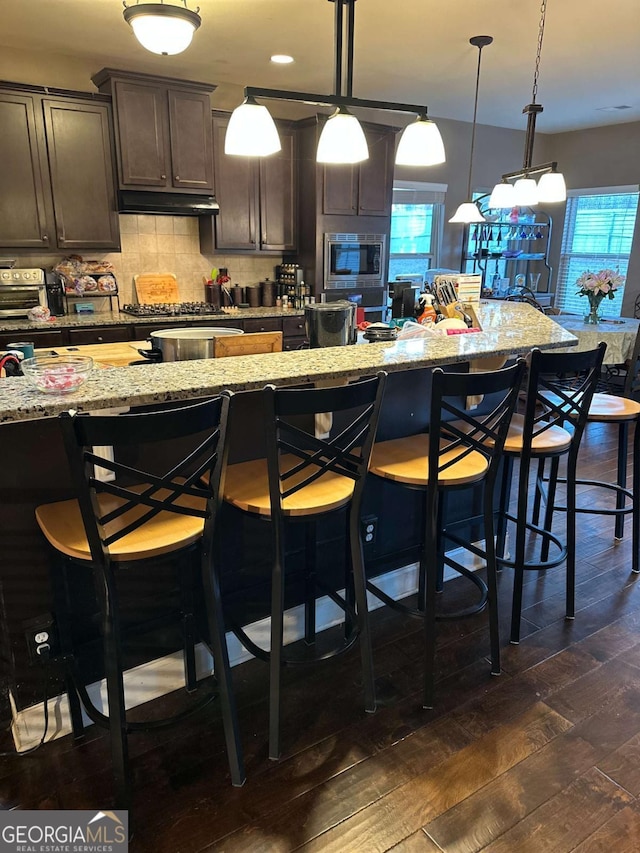 kitchen featuring a kitchen bar, hanging light fixtures, dark brown cabinets, and light stone countertops