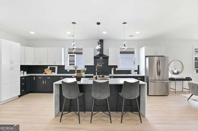 kitchen with white cabinets, an island with sink, wall chimney exhaust hood, freestanding refrigerator, and light countertops