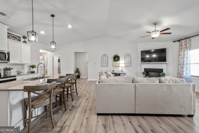 living area with visible vents, a ceiling fan, a glass covered fireplace, light wood-style flooring, and vaulted ceiling