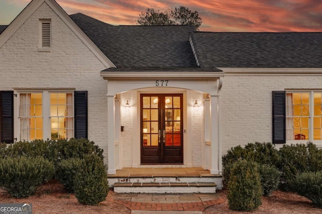 exterior entry at dusk featuring french doors, a shingled roof, and stucco siding