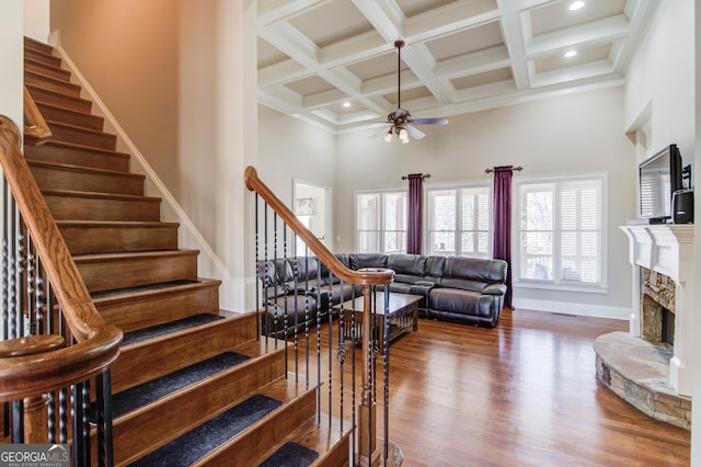 stairs featuring a fireplace, a high ceiling, ceiling fan, wood finished floors, and beamed ceiling