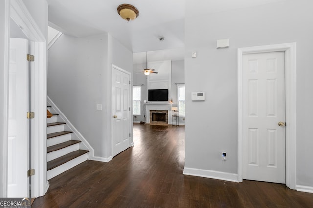 foyer featuring stairs, a ceiling fan, a fireplace with raised hearth, and dark wood-type flooring