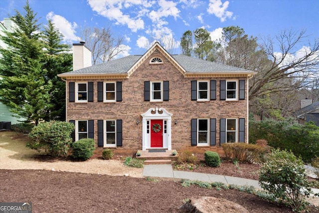 colonial home with roof with shingles, brick siding, a chimney, and central air condition unit