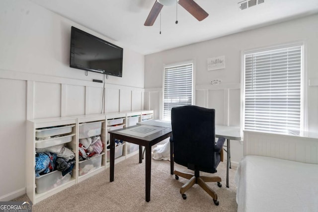 home office featuring ceiling fan, a decorative wall, light carpet, visible vents, and wainscoting