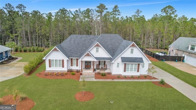 modern farmhouse with roof with shingles, covered porch, a front yard, a view of trees, and driveway