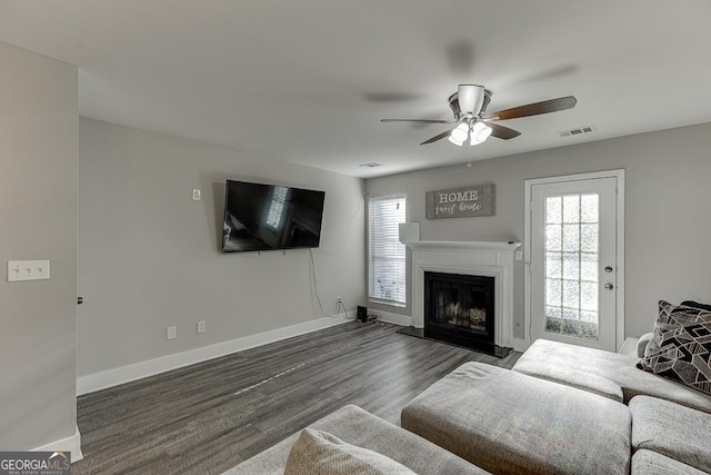 living area with dark wood finished floors, visible vents, a fireplace with raised hearth, a ceiling fan, and baseboards