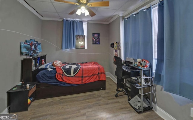 bedroom with ceiling fan, a drop ceiling, wood finished floors, baseboards, and ornamental molding