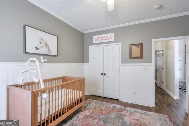 bedroom with a crib, wainscoting, ornamental molding, dark wood-type flooring, and a closet