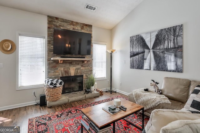 living area with visible vents, vaulted ceiling, a stone fireplace, wood finished floors, and baseboards