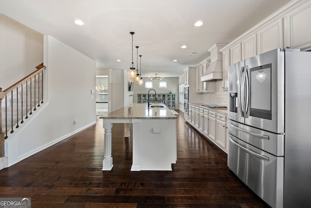 kitchen with premium range hood, white cabinets, hanging light fixtures, appliances with stainless steel finishes, and a center island with sink