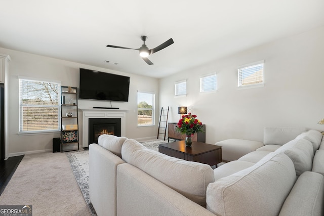 living area featuring a ceiling fan, a glass covered fireplace, light colored carpet, and baseboards