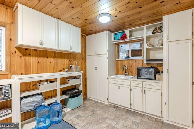 kitchen with wooden ceiling, wooden walls, a sink, white cabinets, and light countertops