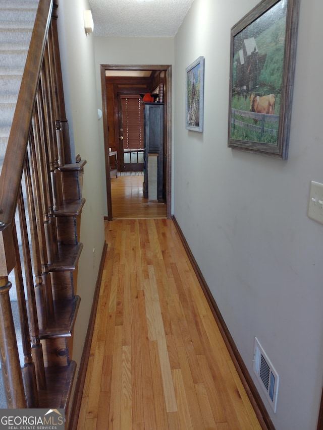 corridor with a textured ceiling, light wood-style flooring, visible vents, and baseboards