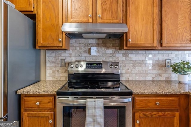 kitchen with light stone counters, under cabinet range hood, stainless steel appliances, brown cabinets, and tasteful backsplash