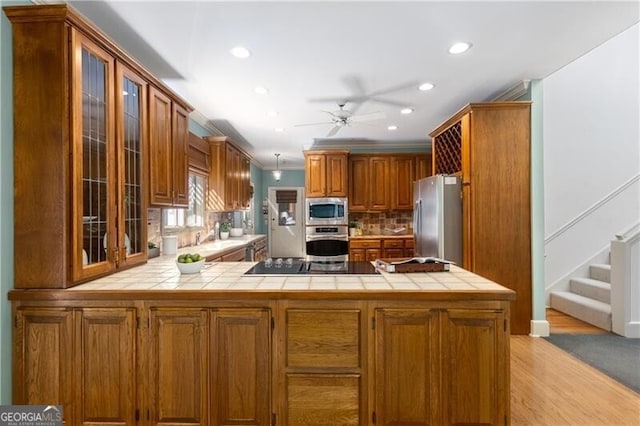 kitchen featuring tile countertops, stainless steel appliances, a peninsula, brown cabinetry, and glass insert cabinets