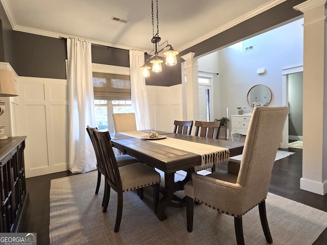 dining room featuring dark wood-style flooring, crown molding, a decorative wall, wainscoting, and ornate columns