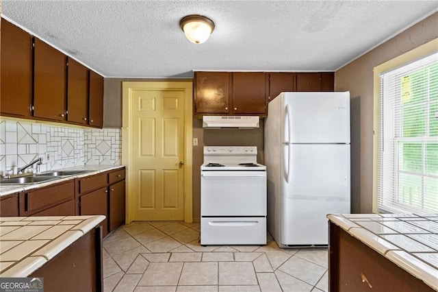 kitchen featuring tile countertops, under cabinet range hood, white appliances, a sink, and tasteful backsplash