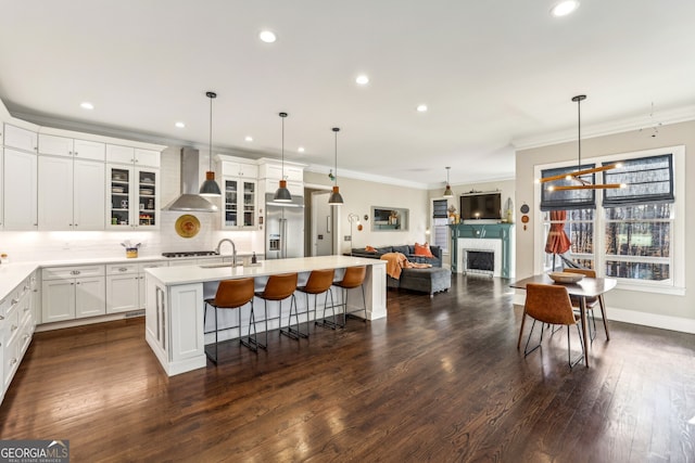 kitchen with built in fridge, a fireplace, light countertops, a kitchen island with sink, and wall chimney exhaust hood