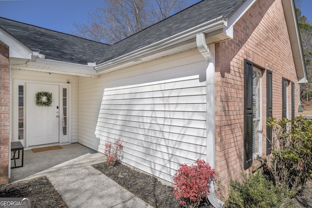 doorway to property with a garage, brick siding, and roof with shingles