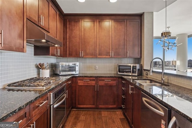 kitchen with stainless steel appliances, dark wood-type flooring, a sink, dark stone countertops, and under cabinet range hood