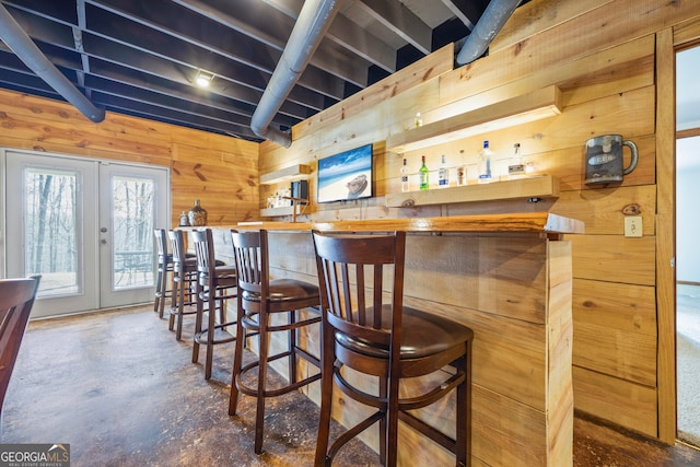 bar featuring french doors, wood walls, a bar, and unfinished concrete floors