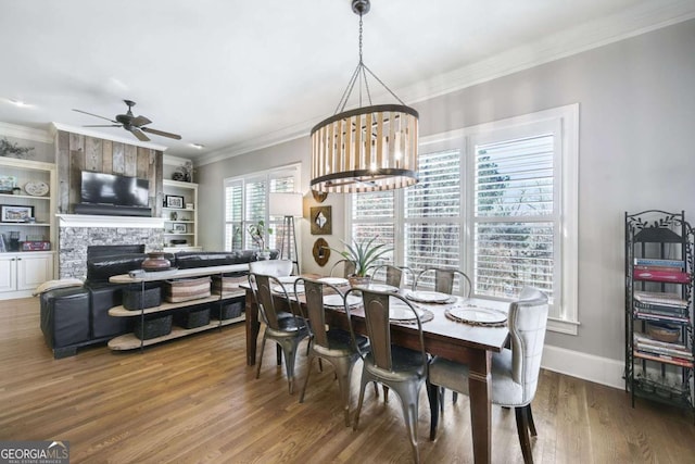 dining area with a large fireplace, ceiling fan with notable chandelier, wood finished floors, baseboards, and crown molding