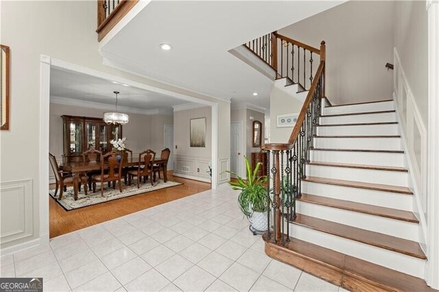 foyer entrance featuring crown molding, a notable chandelier, light tile patterned floors, a decorative wall, and stairs