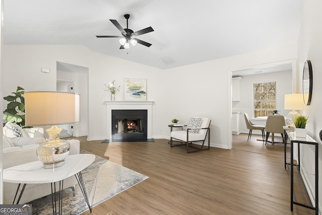 living area featuring lofted ceiling, wood finished floors, a fireplace with flush hearth, visible vents, and a ceiling fan