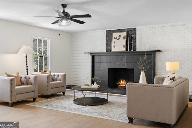 living room with a large fireplace, a ceiling fan, brick wall, wood finished floors, and a textured ceiling