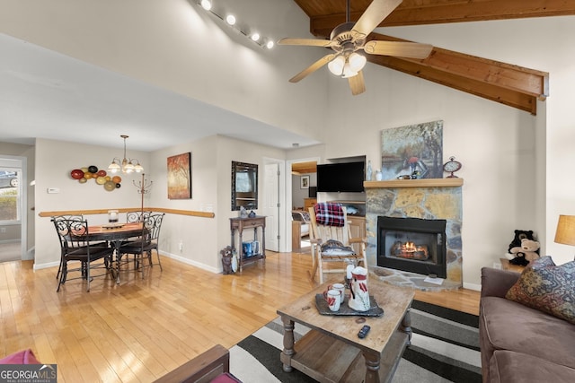 living room with light wood finished floors, baseboards, beamed ceiling, a stone fireplace, and ceiling fan with notable chandelier