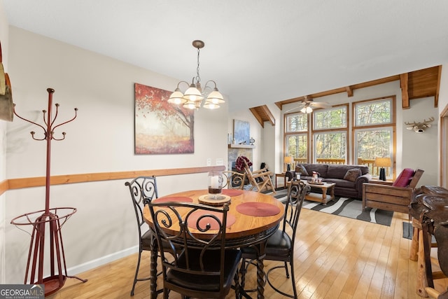 dining space featuring a large fireplace, ceiling fan with notable chandelier, baseboards, light wood-style floors, and beam ceiling