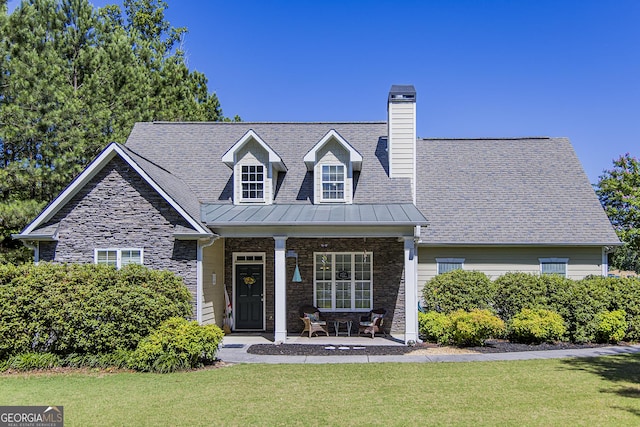 cape cod home with stone siding, a chimney, metal roof, a front lawn, and a porch