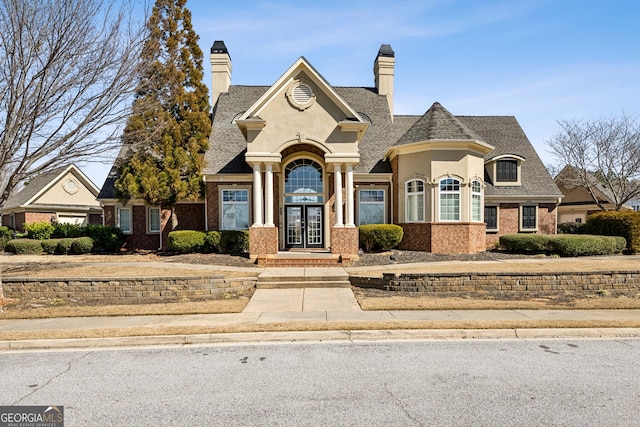 view of front of house featuring brick siding, french doors, roof with shingles, stucco siding, and a chimney