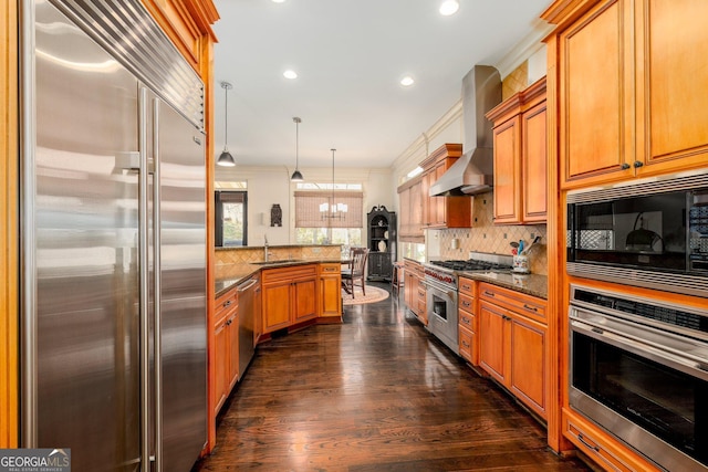 kitchen with a sink, built in appliances, dark stone counters, a peninsula, and wall chimney exhaust hood