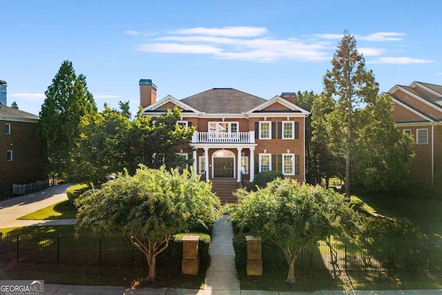 colonial house featuring a balcony, a fenced front yard, a chimney, and brick siding