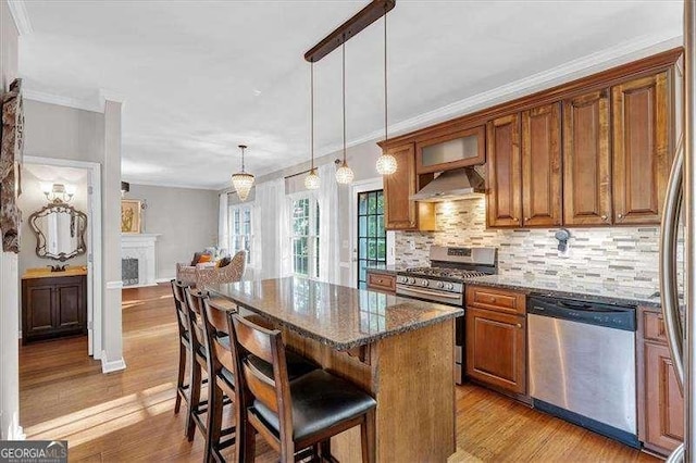 kitchen with stainless steel appliances, light wood-type flooring, wall chimney exhaust hood, brown cabinetry, and dark stone countertops