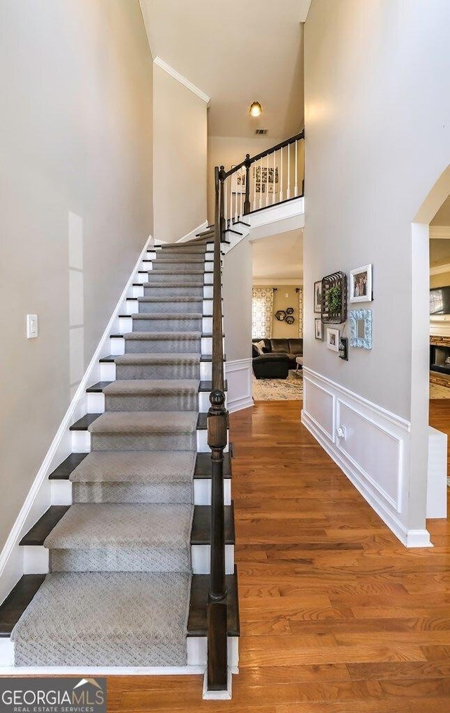 stairway with a decorative wall, a wainscoted wall, a high ceiling, wood finished floors, and ornamental molding