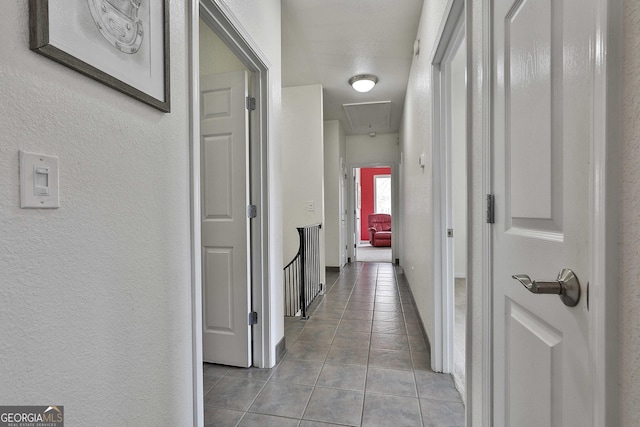 hall featuring a textured wall, light tile patterned flooring, attic access, and baseboards