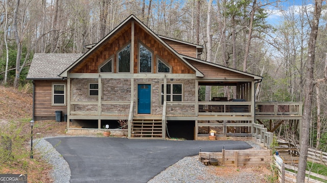 rustic home featuring a shingled roof, a view of trees, stone siding, stairway, and aphalt driveway