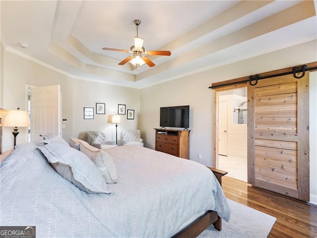 bedroom with a barn door, a ceiling fan, dark wood-style flooring, a tray ceiling, and crown molding